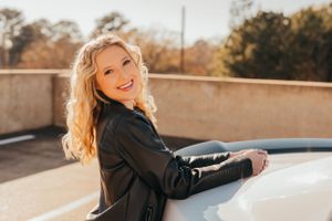 Teen in leather jacket leans on white car during golden hour photoshoot.