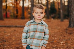 Child in plaid shirt stands among fall foliage in wooded setting during autumn.