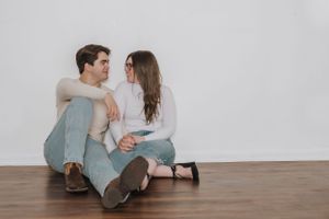 Couple in casual attire sitting close together on wooden floor against white wall.