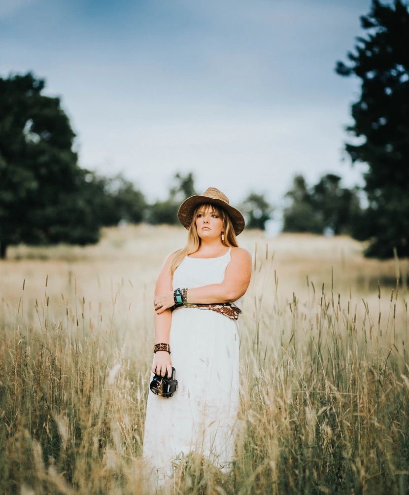 Person in a white dress and straw hat poses in a sunlit field of tall grass.