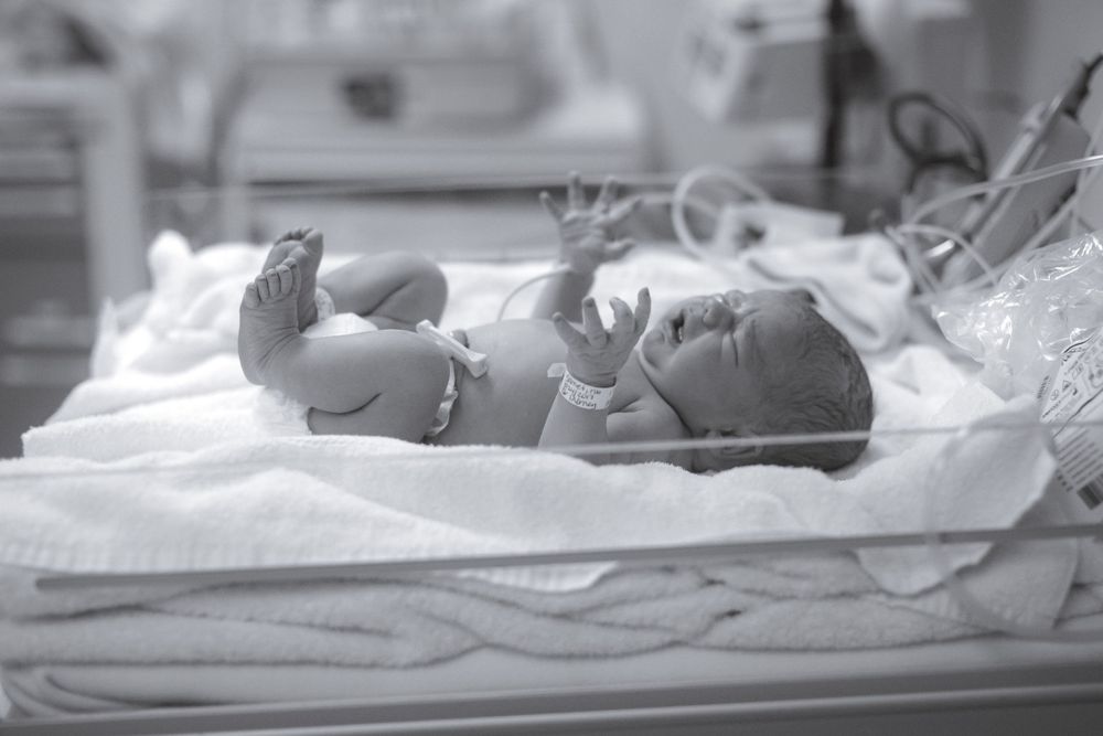 Newborn baby lies on medical examination table under bright hospital lights.