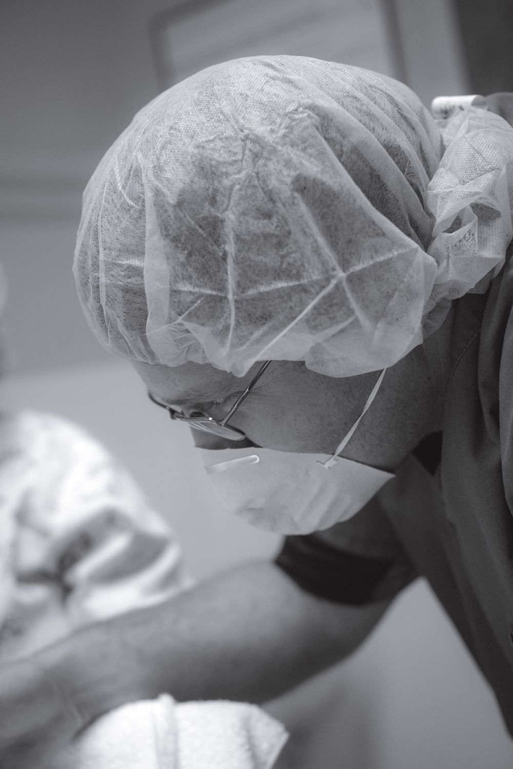 Medical professional in surgical cap and mask leans forward during procedure.