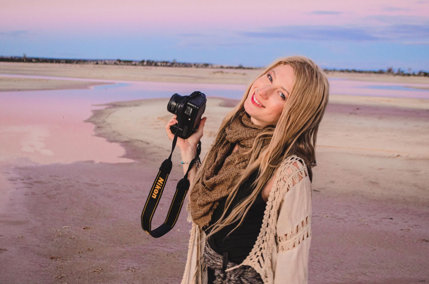 Woman photographer with camera smiling at sunset on beach with pink sky.