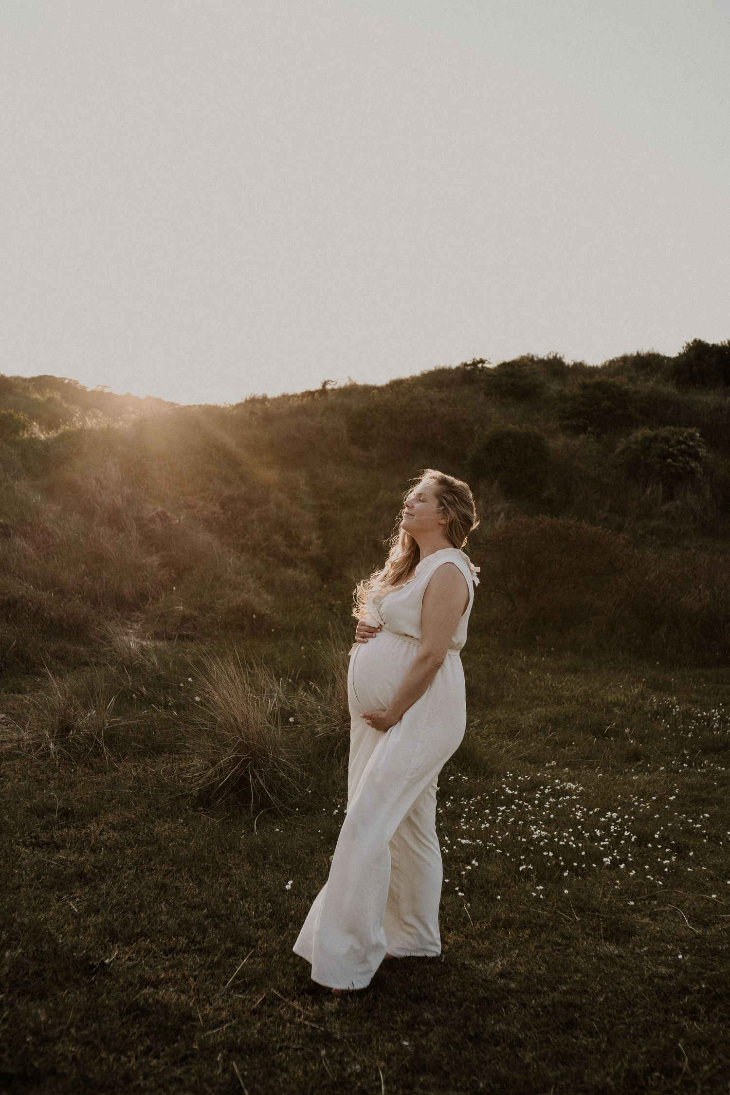 Ethereal maternity photo of woman in white dress at sunset on grassy hillside.