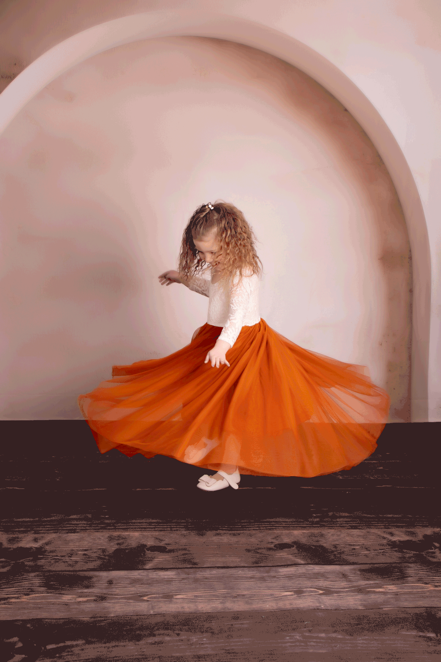 A dancer in a bright orange flowy skirt twirls elegantly against a curved white archway backdrop.