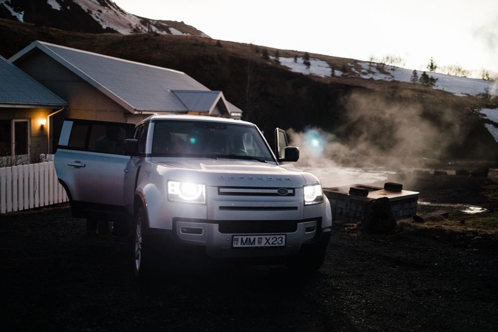 Land Rover Defender with headlights on parked in front of a cabin at dusk with steam rising in cold weather.