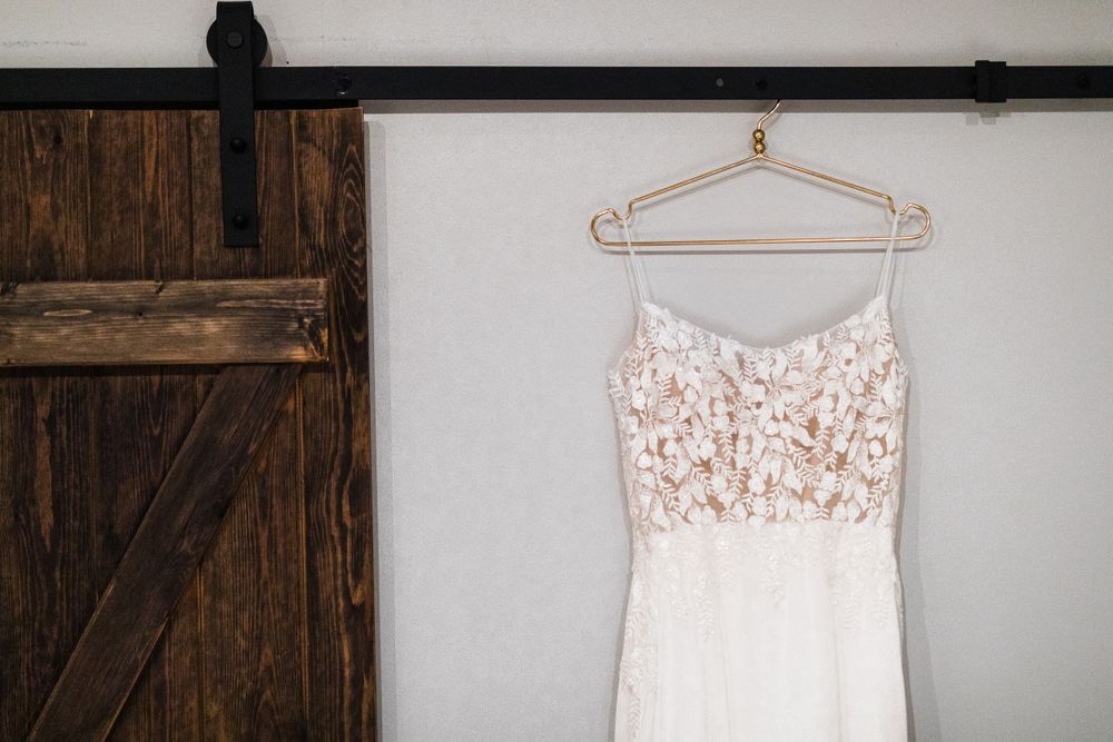 White lace wedding dress hangs on wooden hanger against rustic barn door.