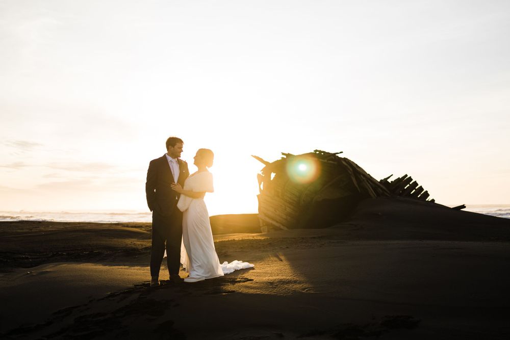 Silhouetted couple in formal attire standing together against dramatic sunset on beach.