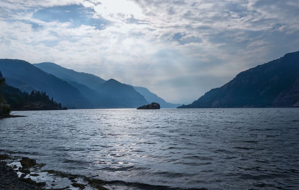 A misty mountain lake stretches between forested slopes under cloudy skies.