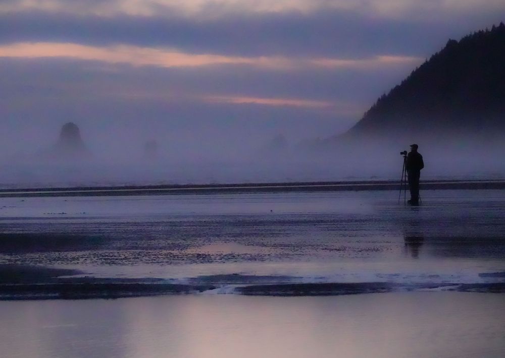 Silhouetted figure photographs misty Cannon Beach at twilight with dramatic coastal formations.