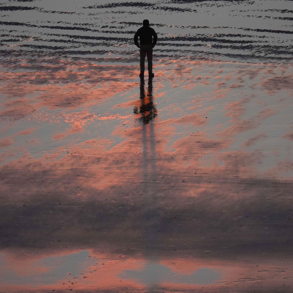 Solitary figure reflected in wet sand during vibrant orange sunset at the beach.