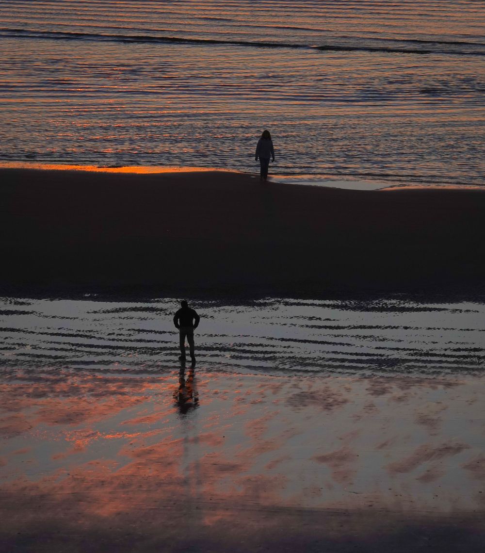 Two silhouettes on beach at sunset create reflections in wet sand with orange sky.