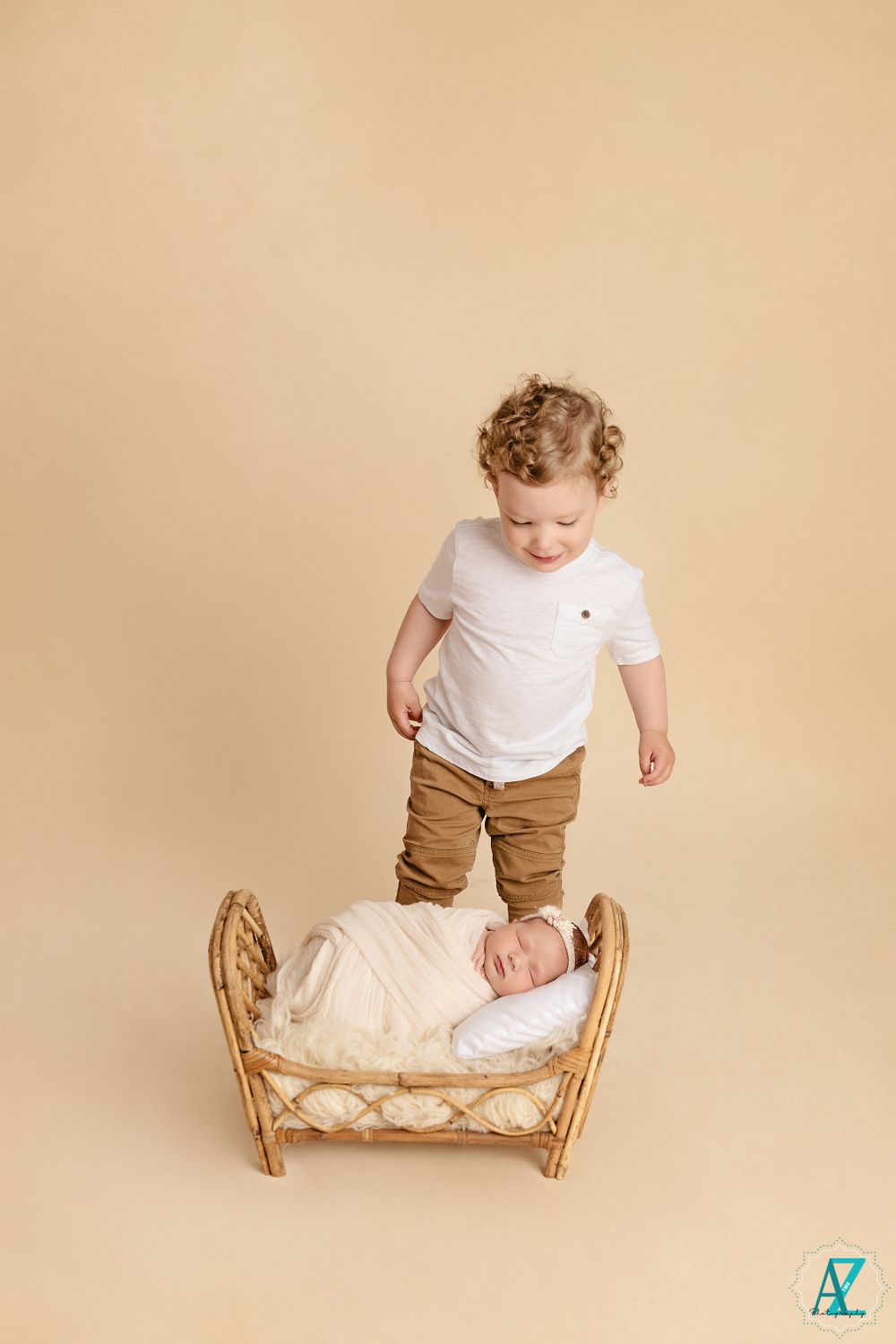 A toddler stands beside a small wicker bed containing a swaddled newborn on a beige background.