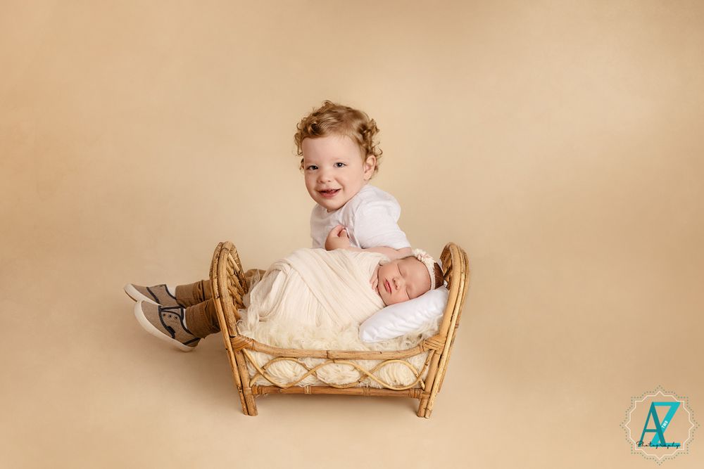 A heartwarming sibling moment captured beside a decorative wicker bed on neutral backdrop.