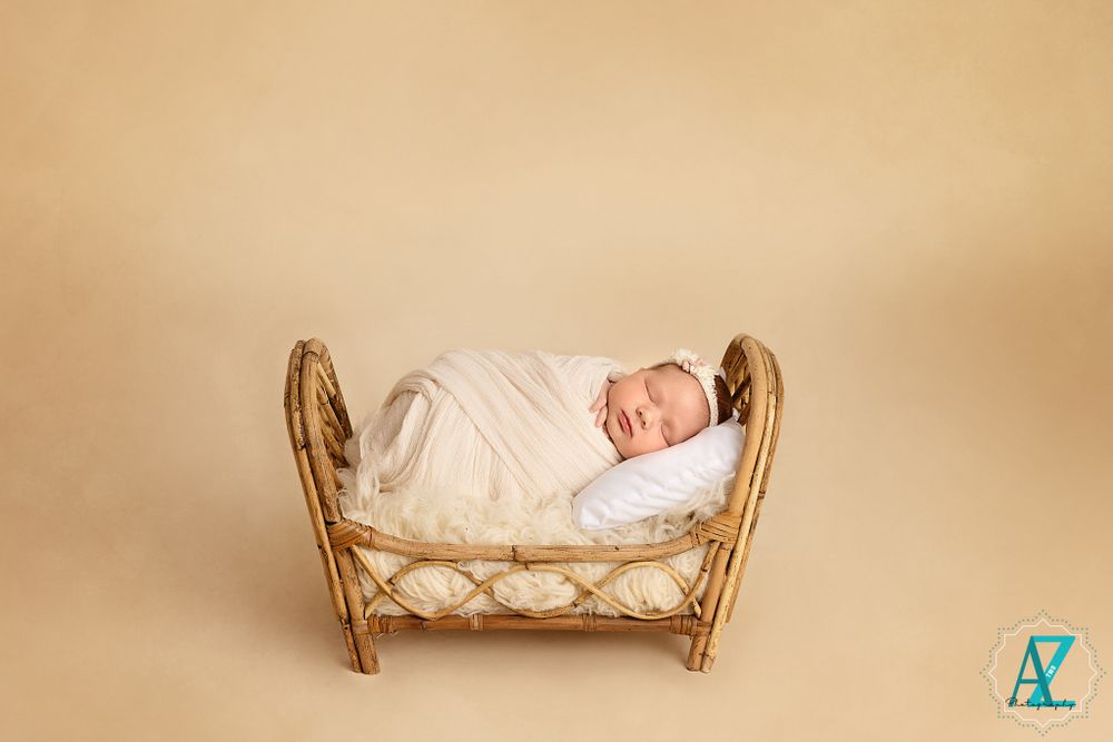 Sleeping newborn swaddled in white lying in a woven rattan bed against beige background.