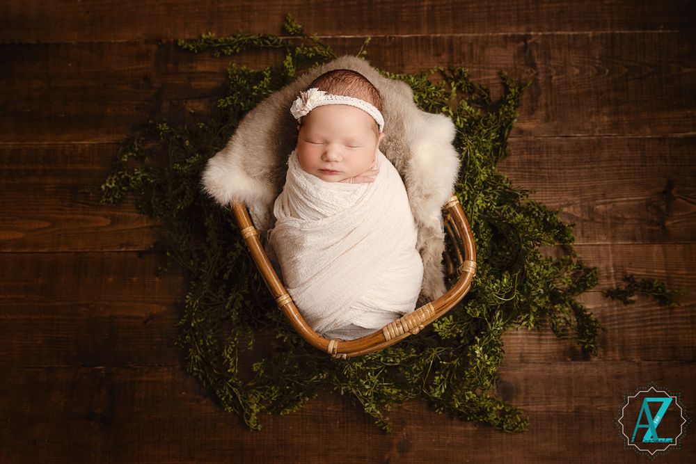 Newborn wrapped in white blanket sleeping in basket surrounded by green moss on wooden backdrop.