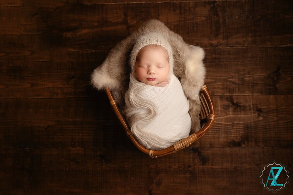 Sleeping baby in white wrap nestled in wooden basket with fur lining against dark wood background.