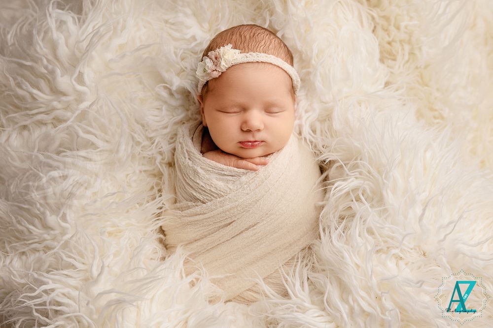 Peaceful newborn wrapped in cream fabric resting on fluffy white fur with delicate headband.