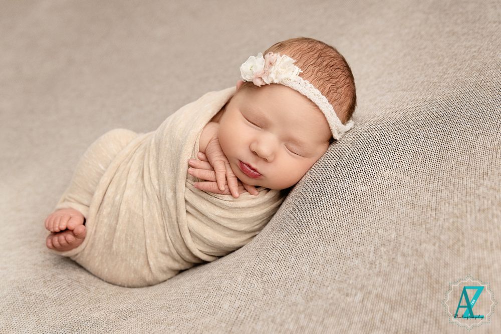 Swaddled infant wearing lace headband sleeping peacefully on soft gray fabric.