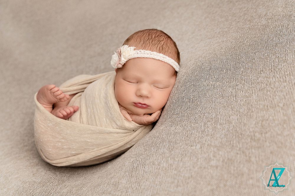 Sleeping newborn in cream wrap with delicate headband on textured gray background.