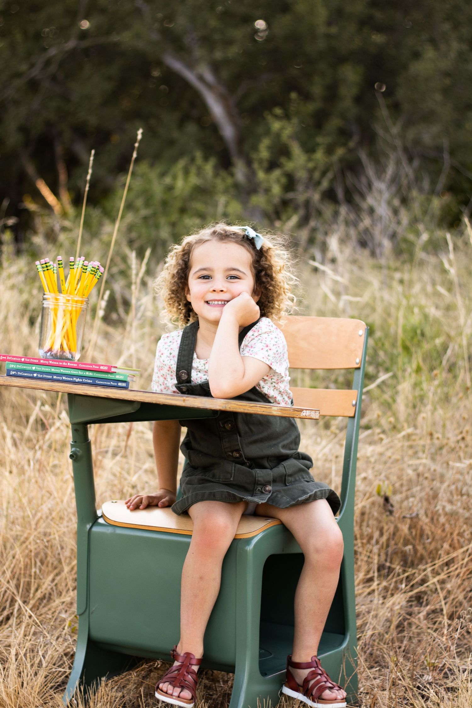 Young girl sitting and posing playfully on vintage green school desk in natural outdoor setting.