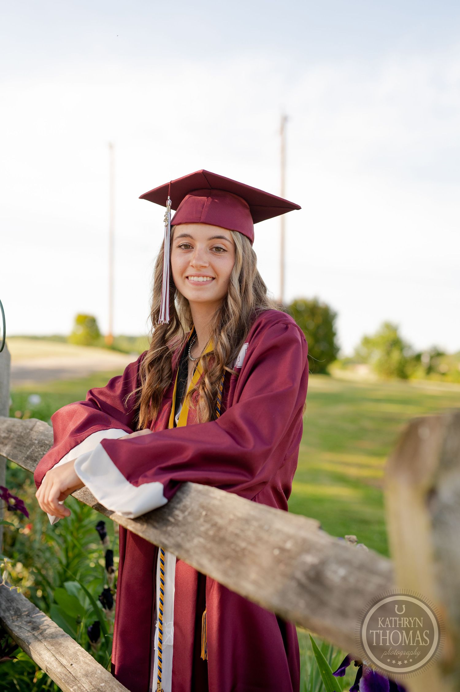 Graduate in burgundy cap and gown leans against rustic wooden fence in outdoor setting during golden hour.