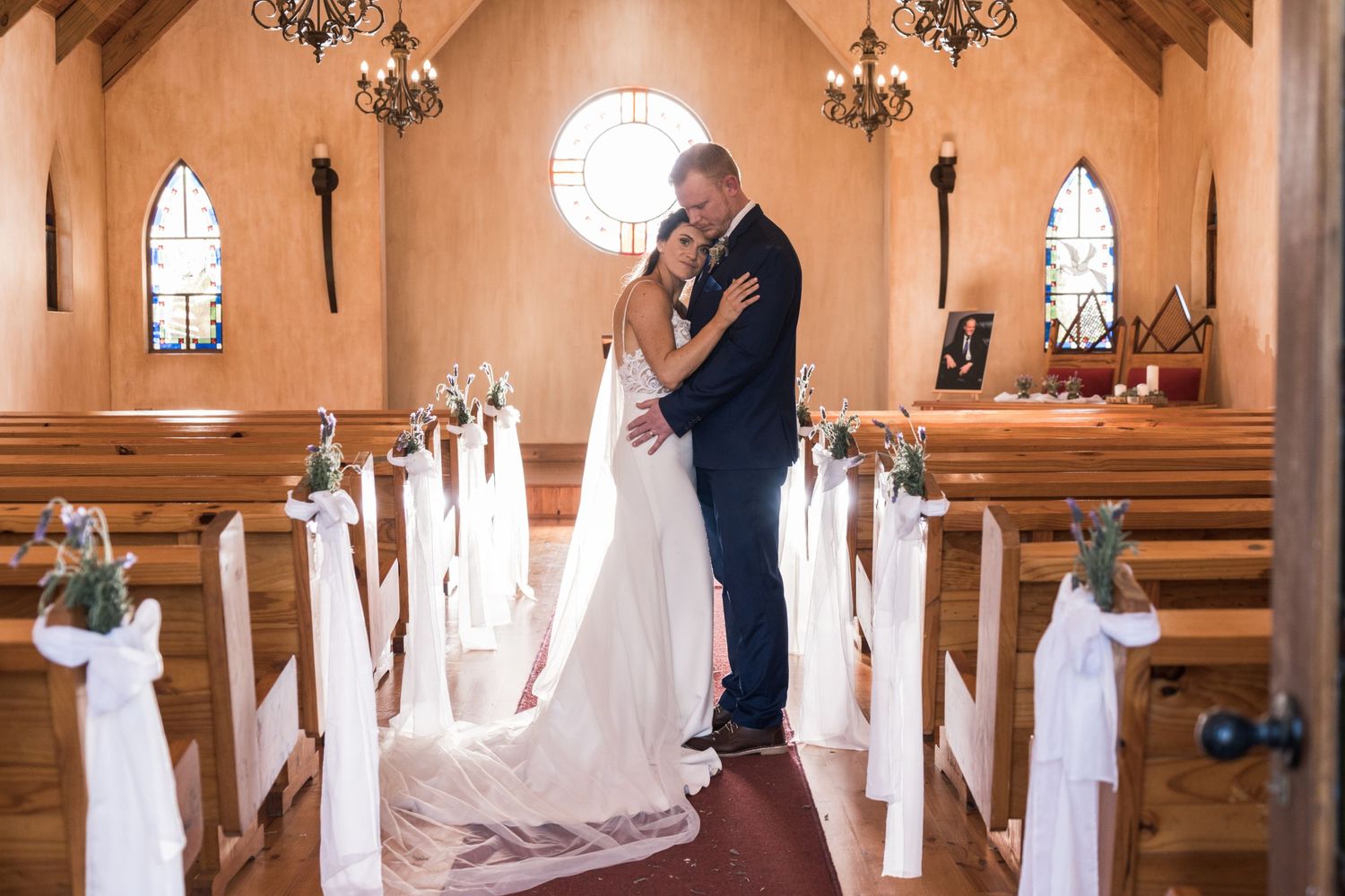 Bride and groom embracing at the altar of a charming chapel with white ribbon-decorated pews.