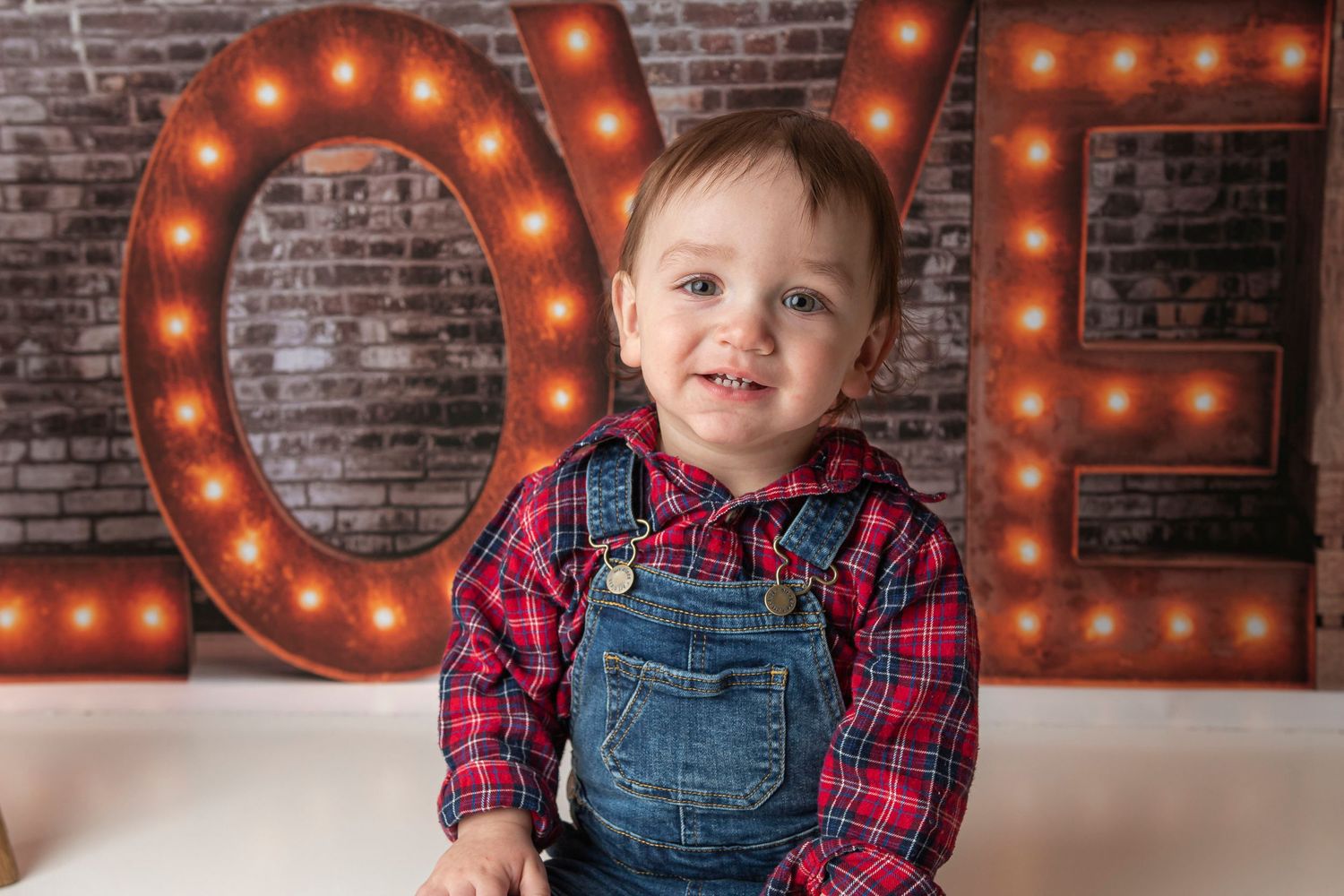 Child in plaid shirt and overalls poses in front of rustic brick wall with glowing LOVE sign.