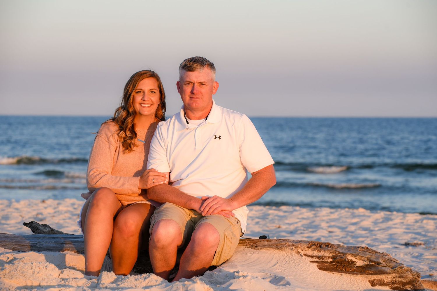 A couple sits together on white sand beach at sunset with ocean waves behind them.