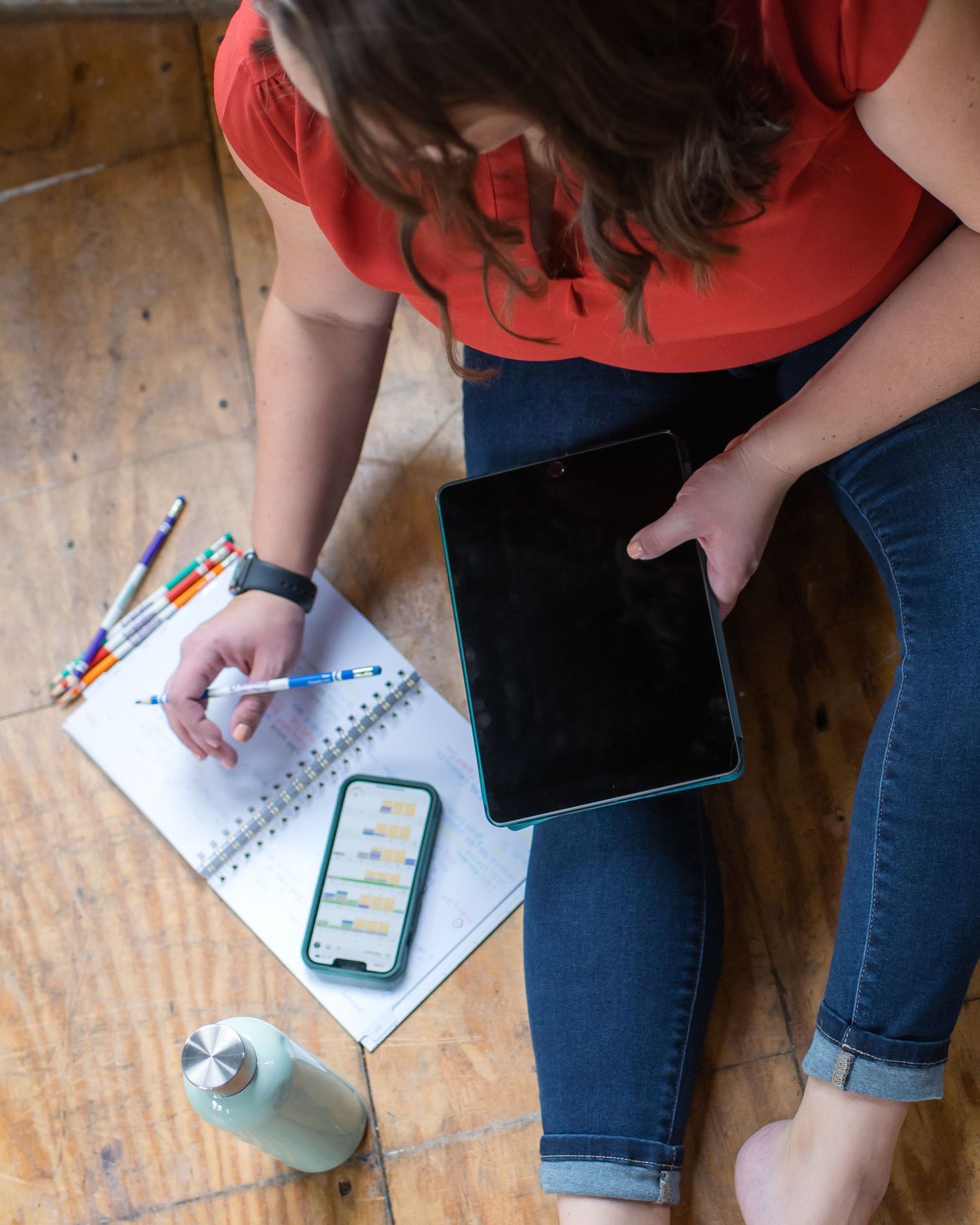 Overhead view of someone working with a tablet and notebook on wooden floor.