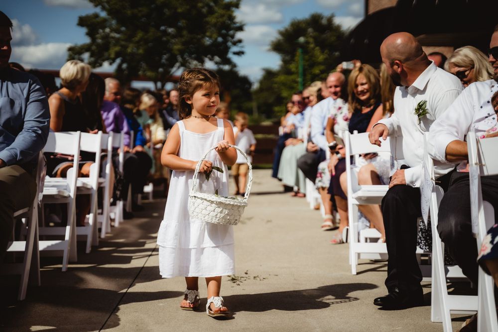Young attendant in white dress walks down outdoor wedding aisle carrying flowers.