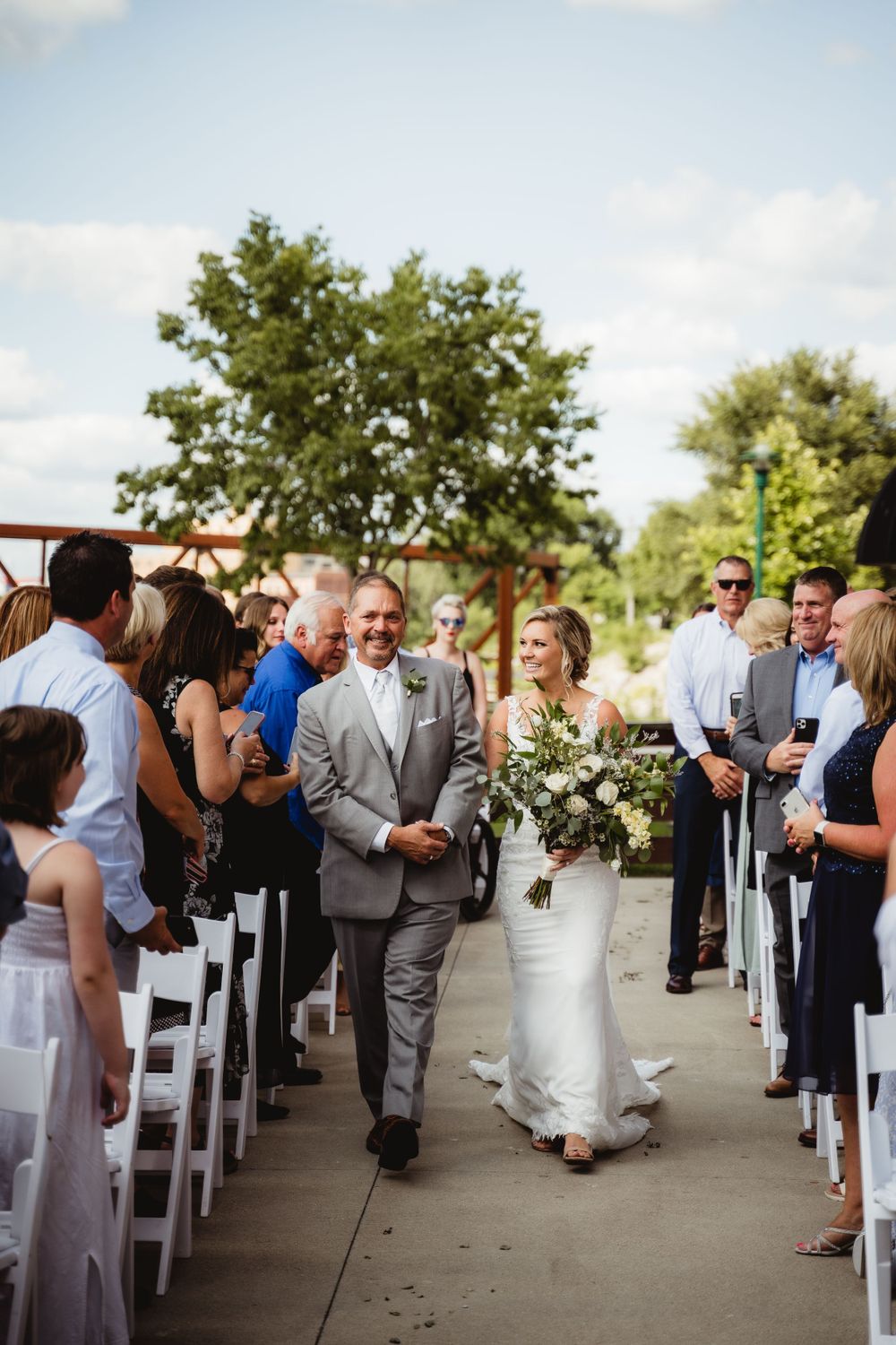 Couple walks down aisle after outdoor wedding ceremony with white chairs and floral arch.