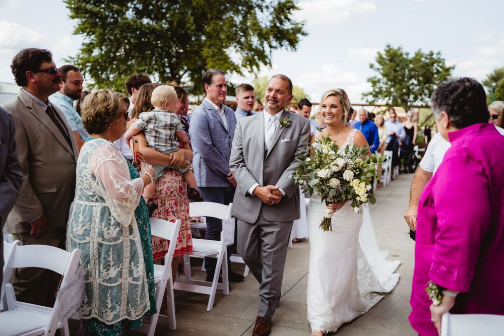 Wedding guests line an outdoor ceremony aisle as a couple walks past in formal attire.