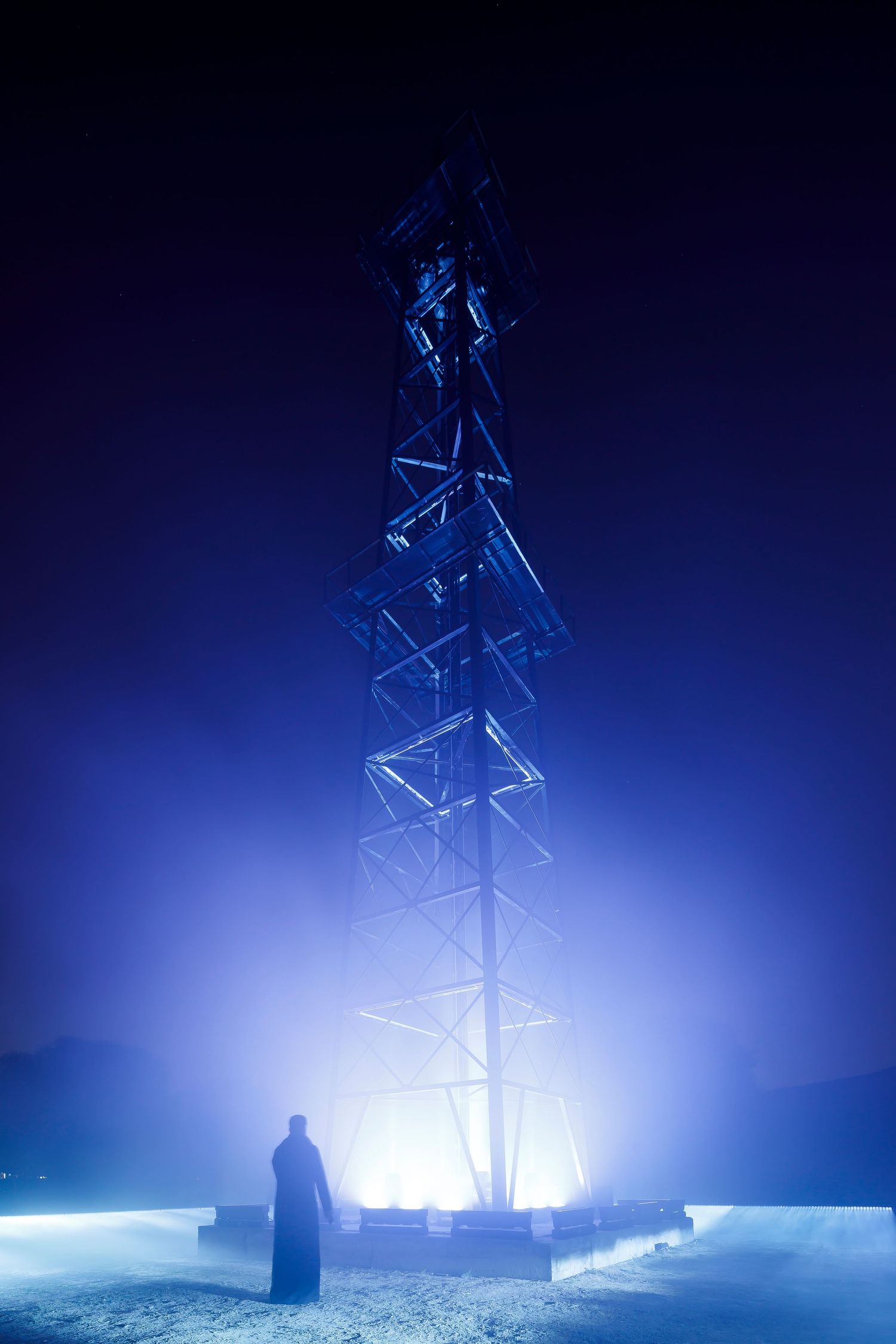 Blue-lit communications tower in foggy night with silhouetted figure standing below.
