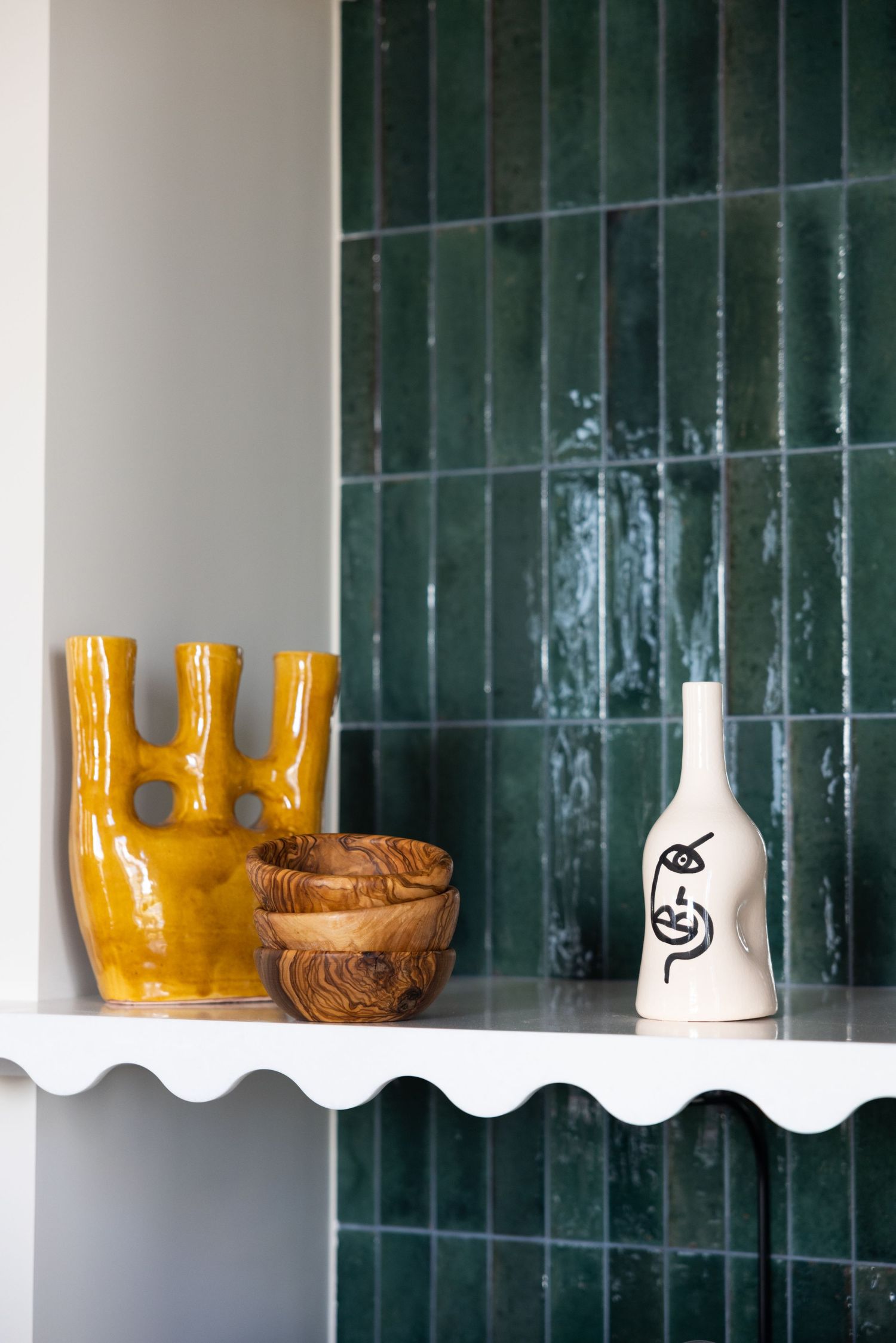 Decorative yellow ceramic vase and white bottle displayed on shelf against green tiled wall.