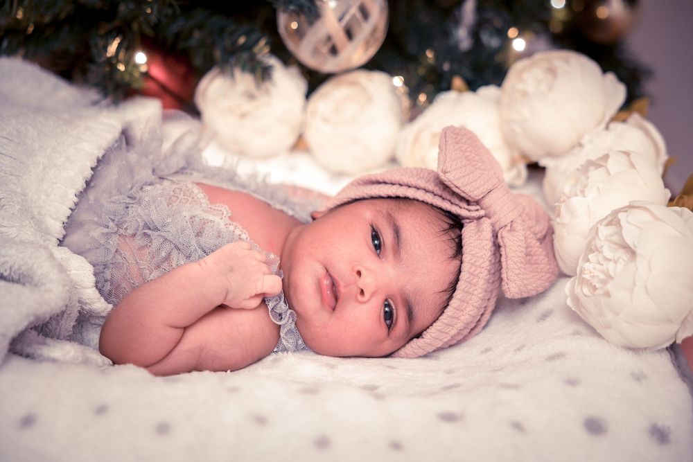 Baby wearing pink knit hat lies on white blanket with decorative holiday lights and ornaments.