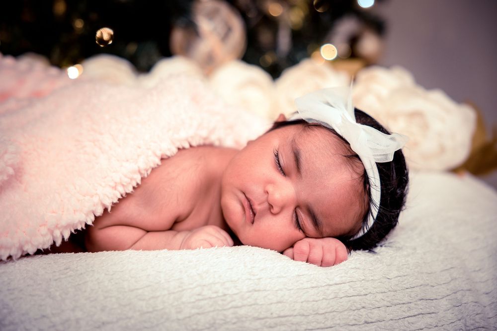 Peaceful sleeping newborn wearing white headband on textured white blanket with bokeh lights in background.