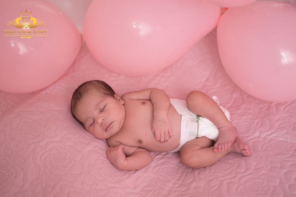 Sleeping newborn baby rests peacefully on pink bedding surrounded by pink balloons in a studio setting.