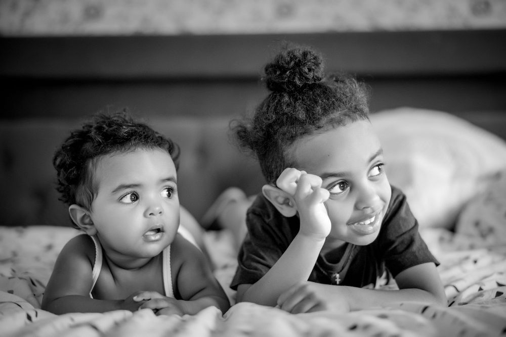 Two children lying on bed making playful expressions in black and white photo.