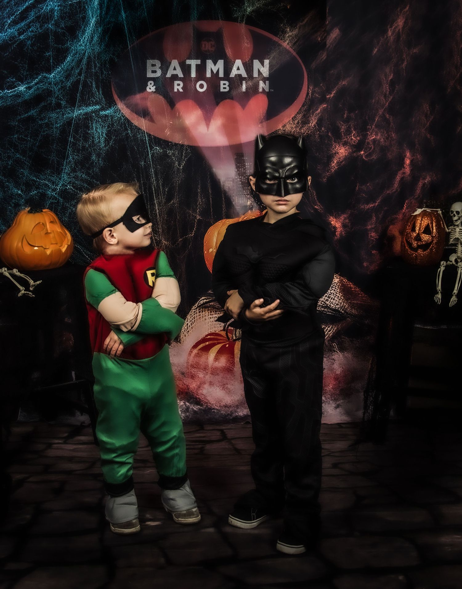 Dark Halloween scene featuring Batman and Robin costumed figures with jack-o-lanterns and spooky lighting.
