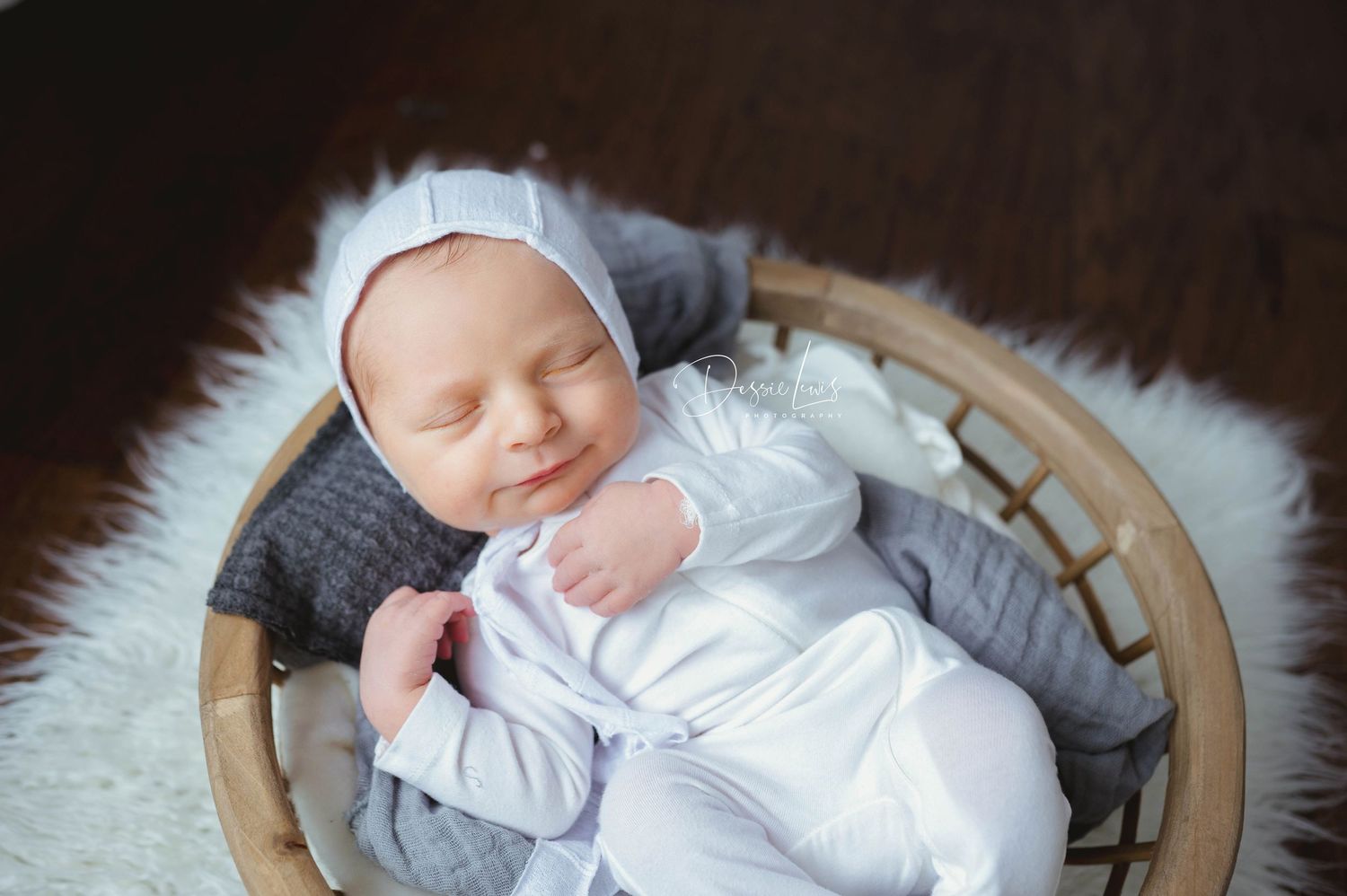 Sleeping newborn in white outfit lying in wooden basket with soft gray padding.
