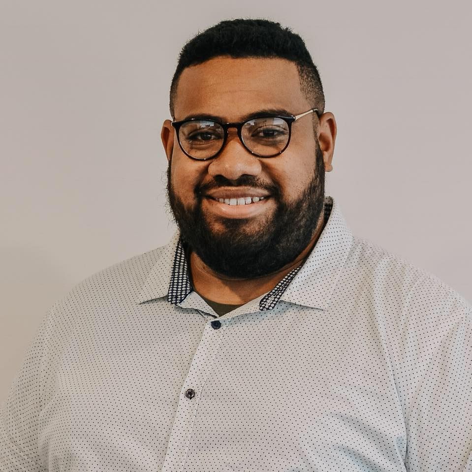 Professional headshot of smiling bearded man wearing glasses and light blue patterned shirt.