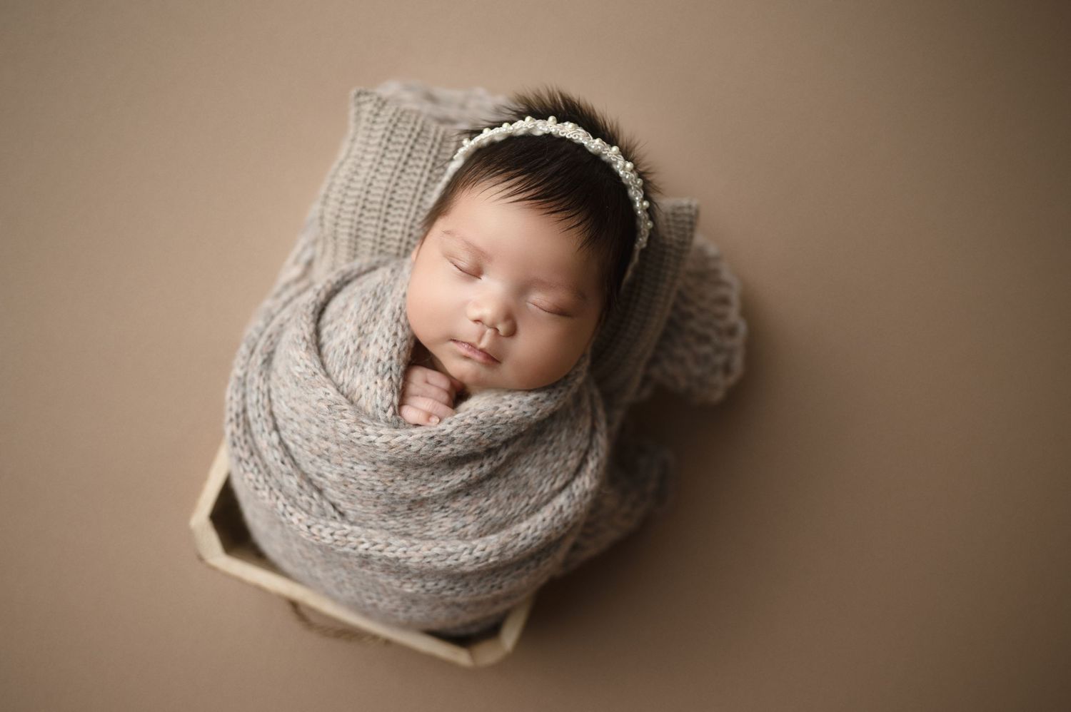 Newborn sleeping peacefully wrapped in gray knit blanket on neutral background.