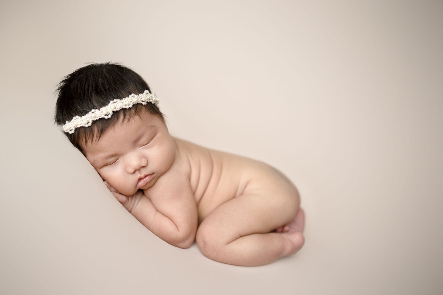 Peaceful newborn resting on white fabric with delicate lace headband in neutral studio setting.