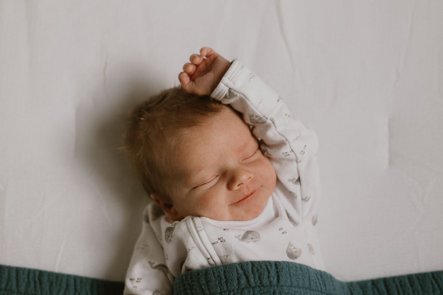 A sleeping newborn baby wearing white stretches their tiny hand upward.