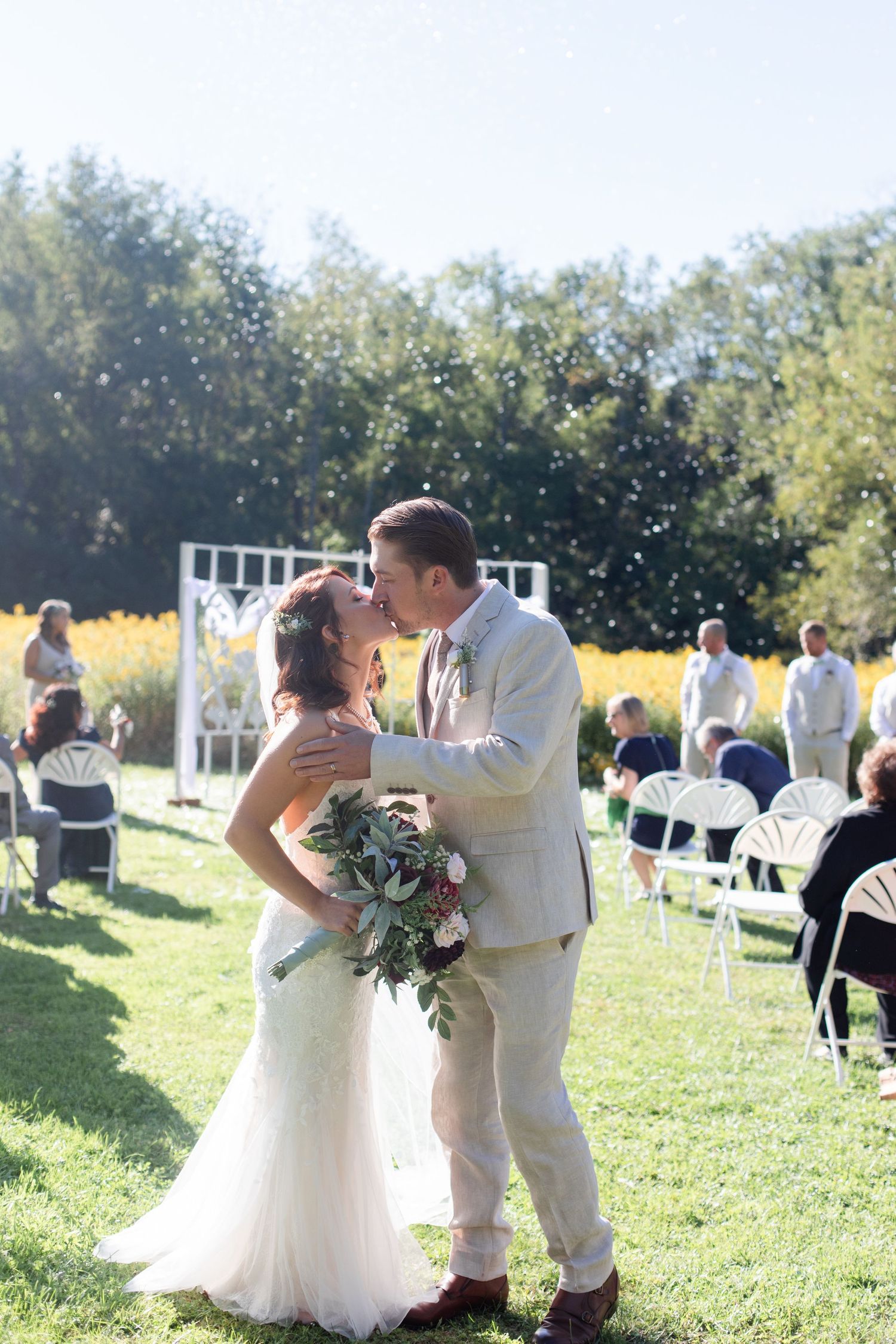 A couple celebrates their wedding moment in a sun-drenched field with guests looking on.