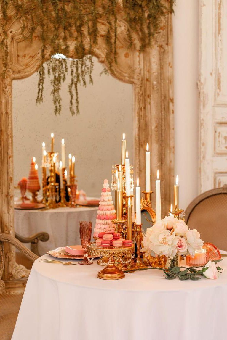 Elegant table setting with gold candlesticks, pink desserts, and floral arrangements under draped greenery.