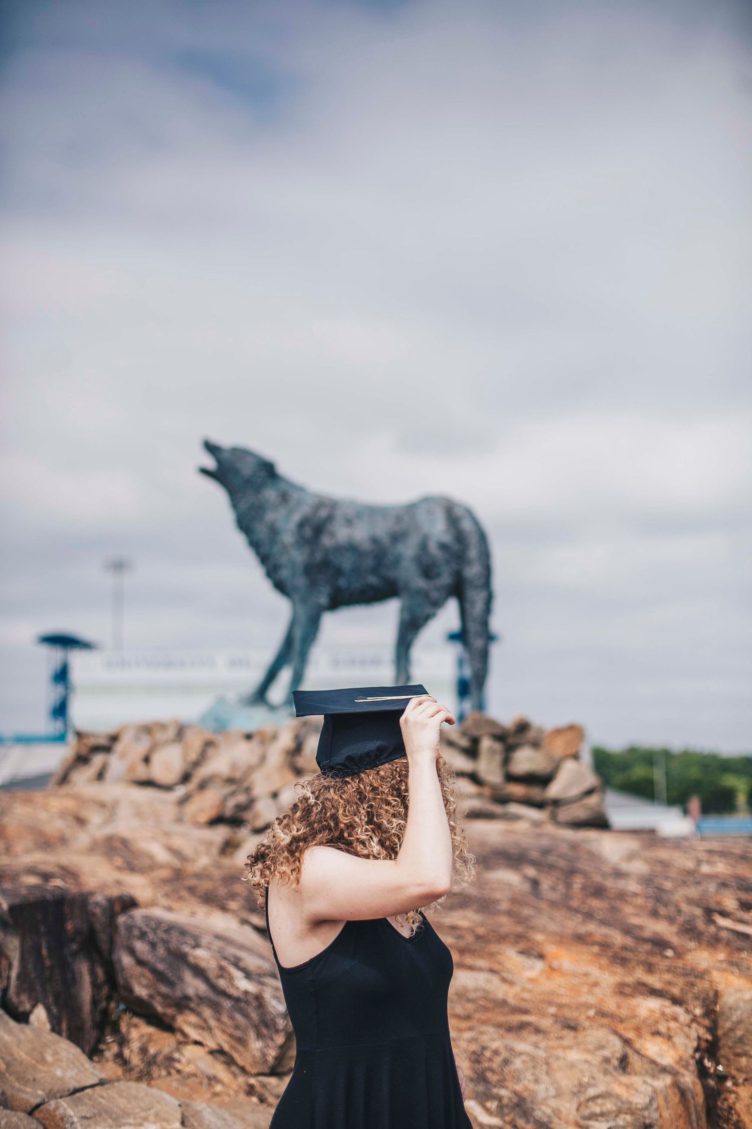 Curly-haired graduate hiding face behind black mortarboard cap near howling wolf sculpture.