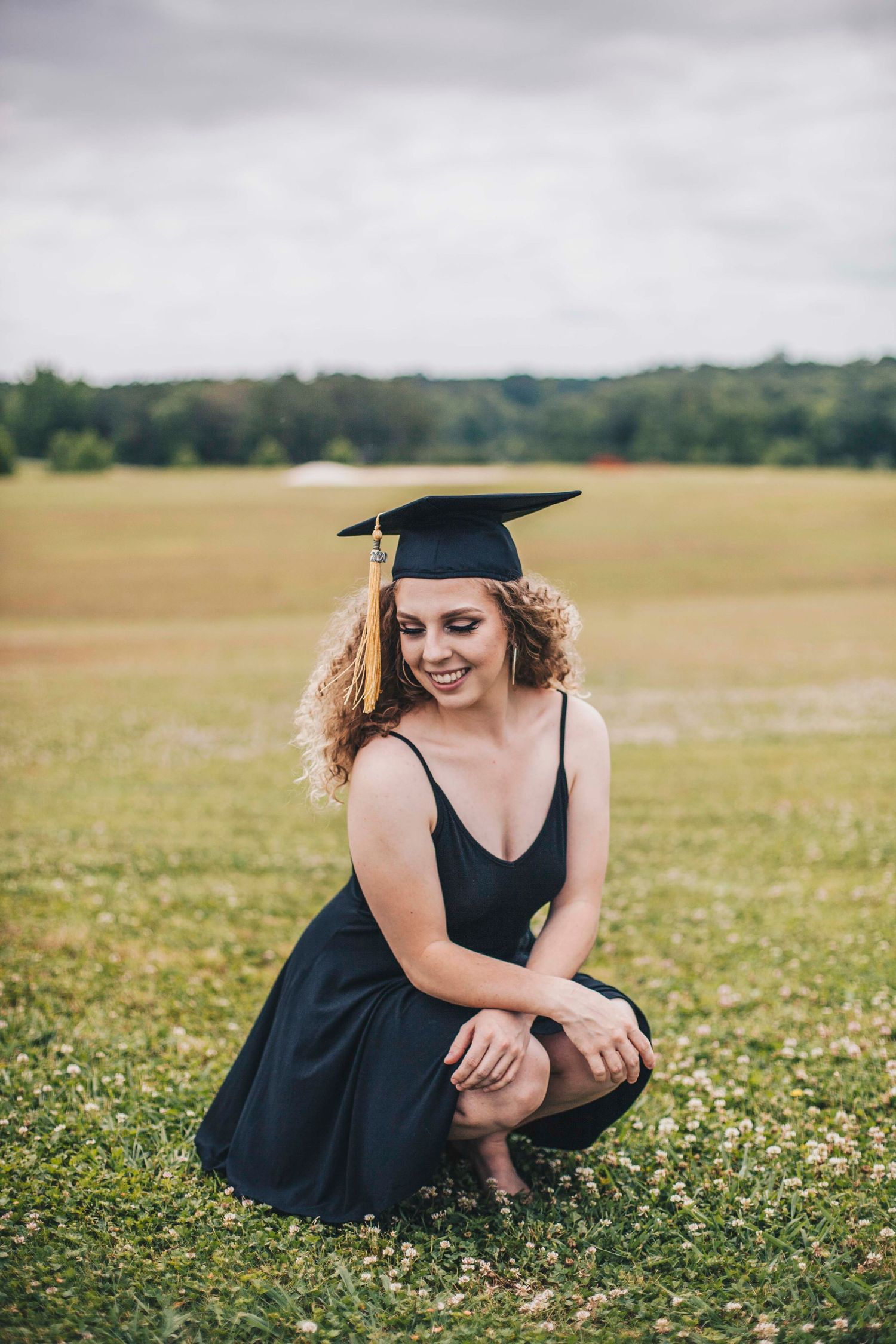 Smiling female graduate in black dress and mortarboard cap crouches on green grass field.