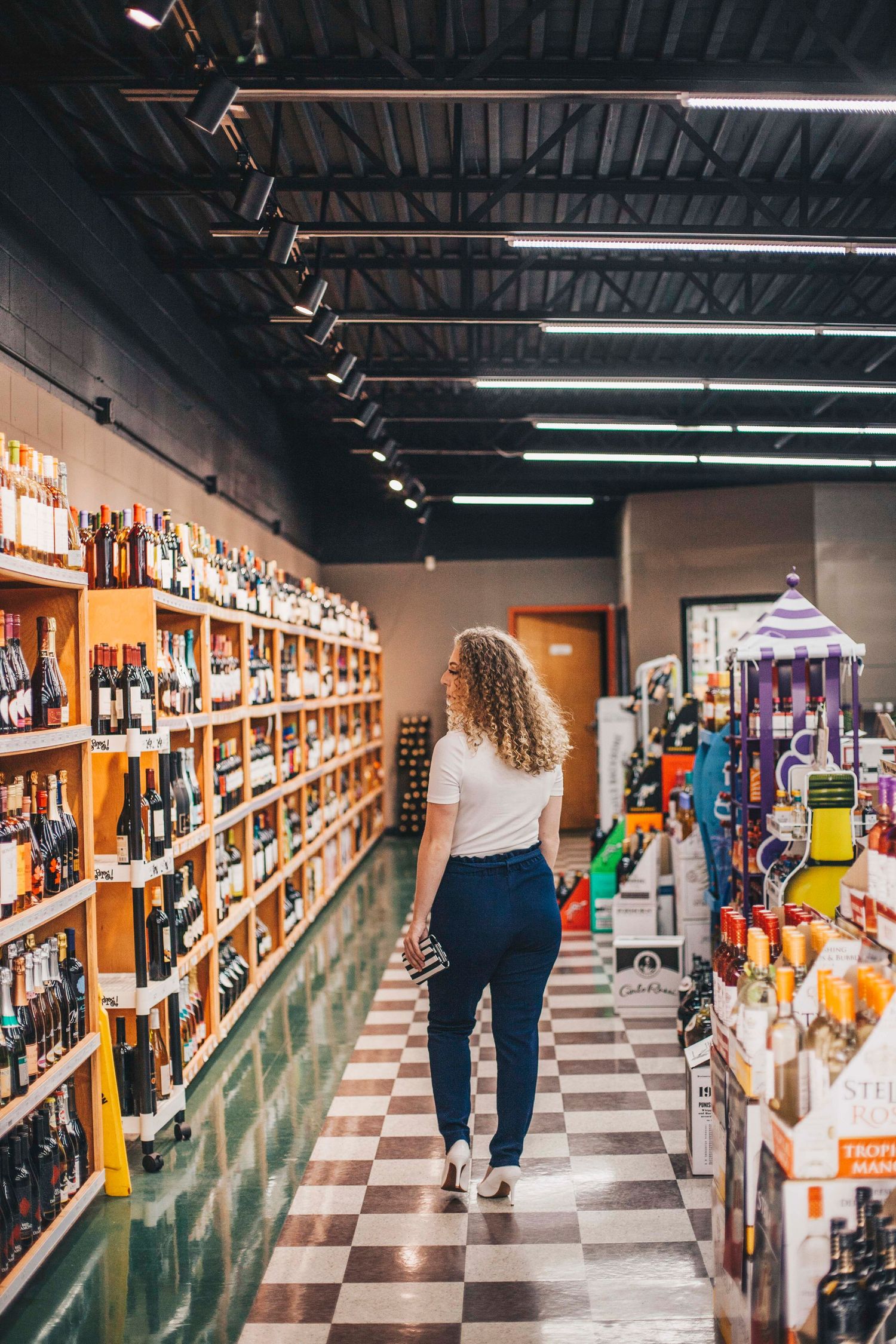 Young curly-haired woman walks through wine store aisle with checkered floor and stocked shelves.
